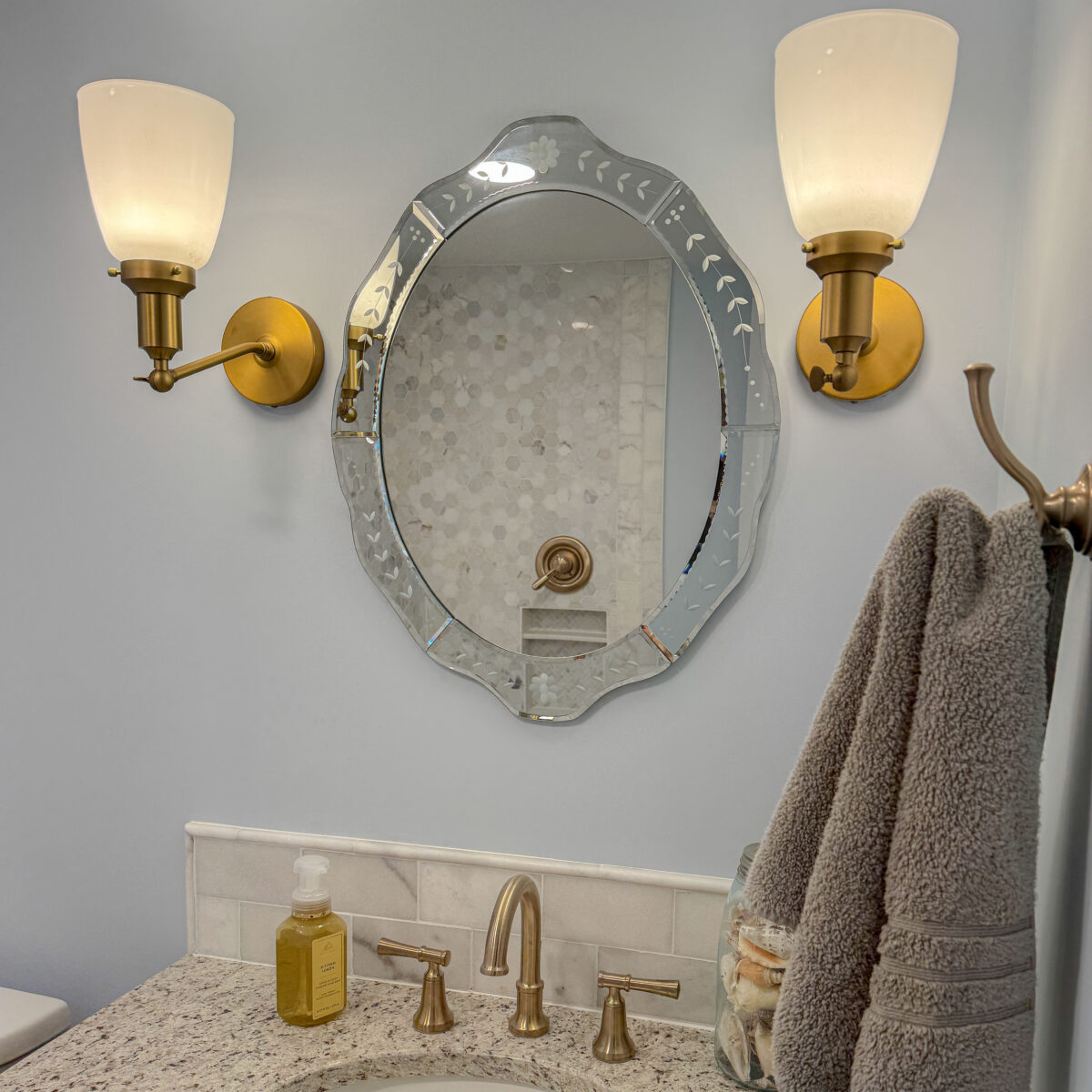 a silver mirror hanging on a wall with brass colored sconces, a brass colored towel hook, and brass faucet on a tan speckled vanity top