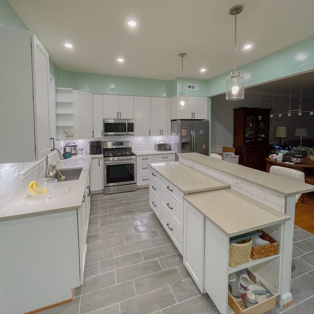 A kitchen with grey tile flooring, white cabinets, a cool toned tan counter, stainless steel appliances, hanging lights over bar seating on the island, and recessed lighting overhead with a light teal colored paint on the walls