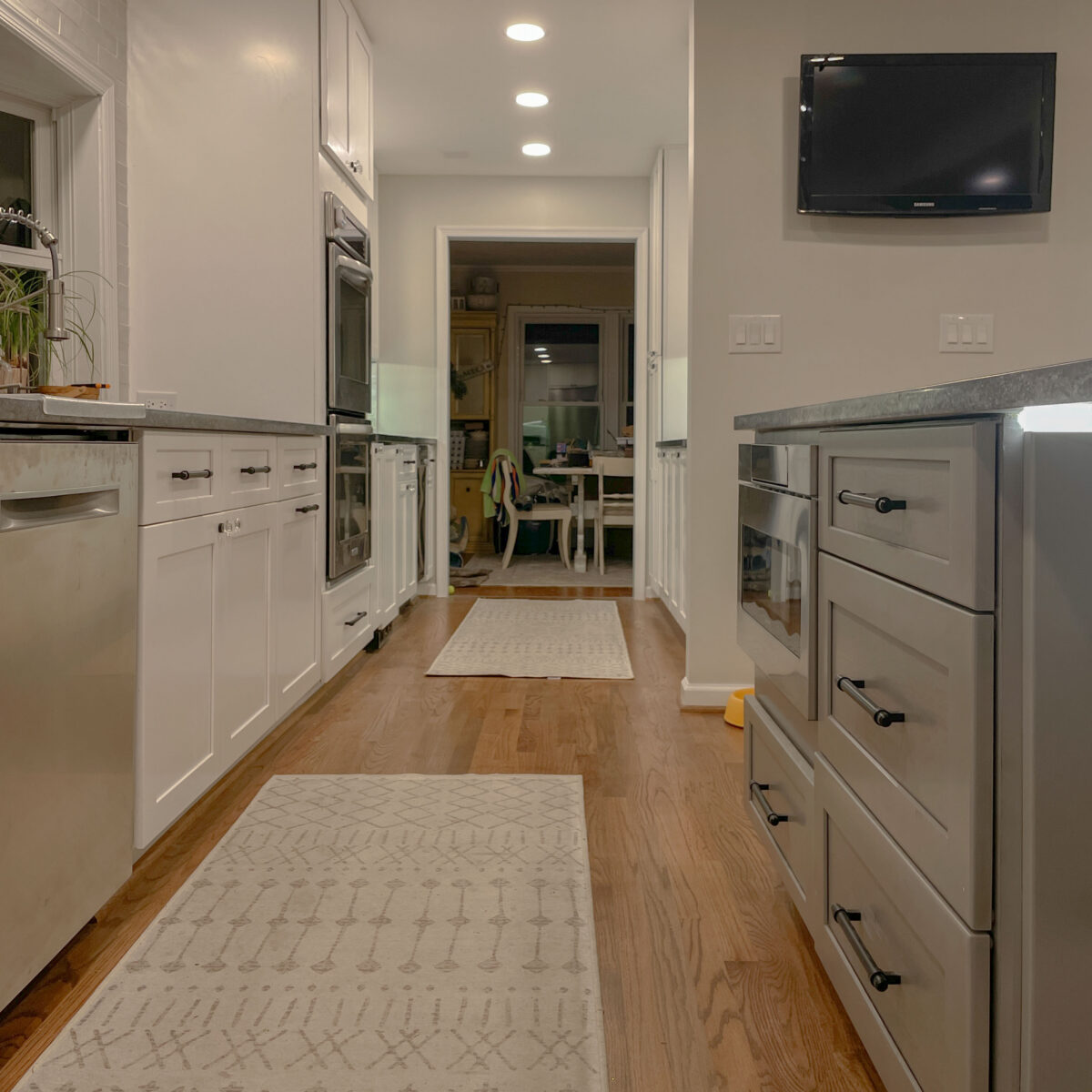 low view of a kitchen with hardwood floors, tan rugs, white cabinets with darker nickel hardware, grey colored counters and a tv mounted on the wall